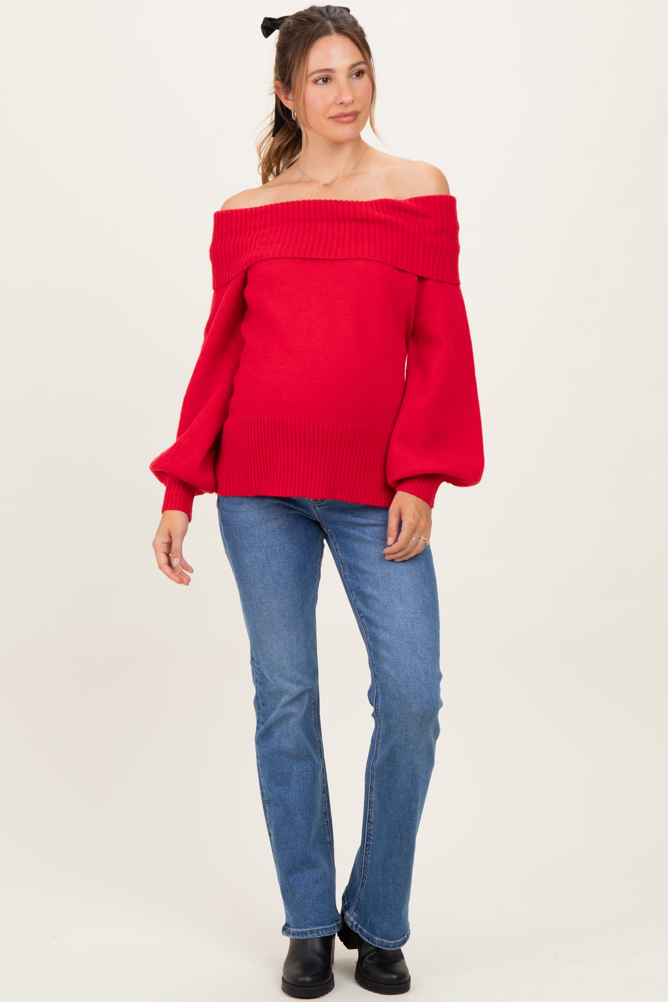 woman wearing a red off-the-shoulder maternity sweater with puff sleeves and a baby bump visible, paired with denim jeans