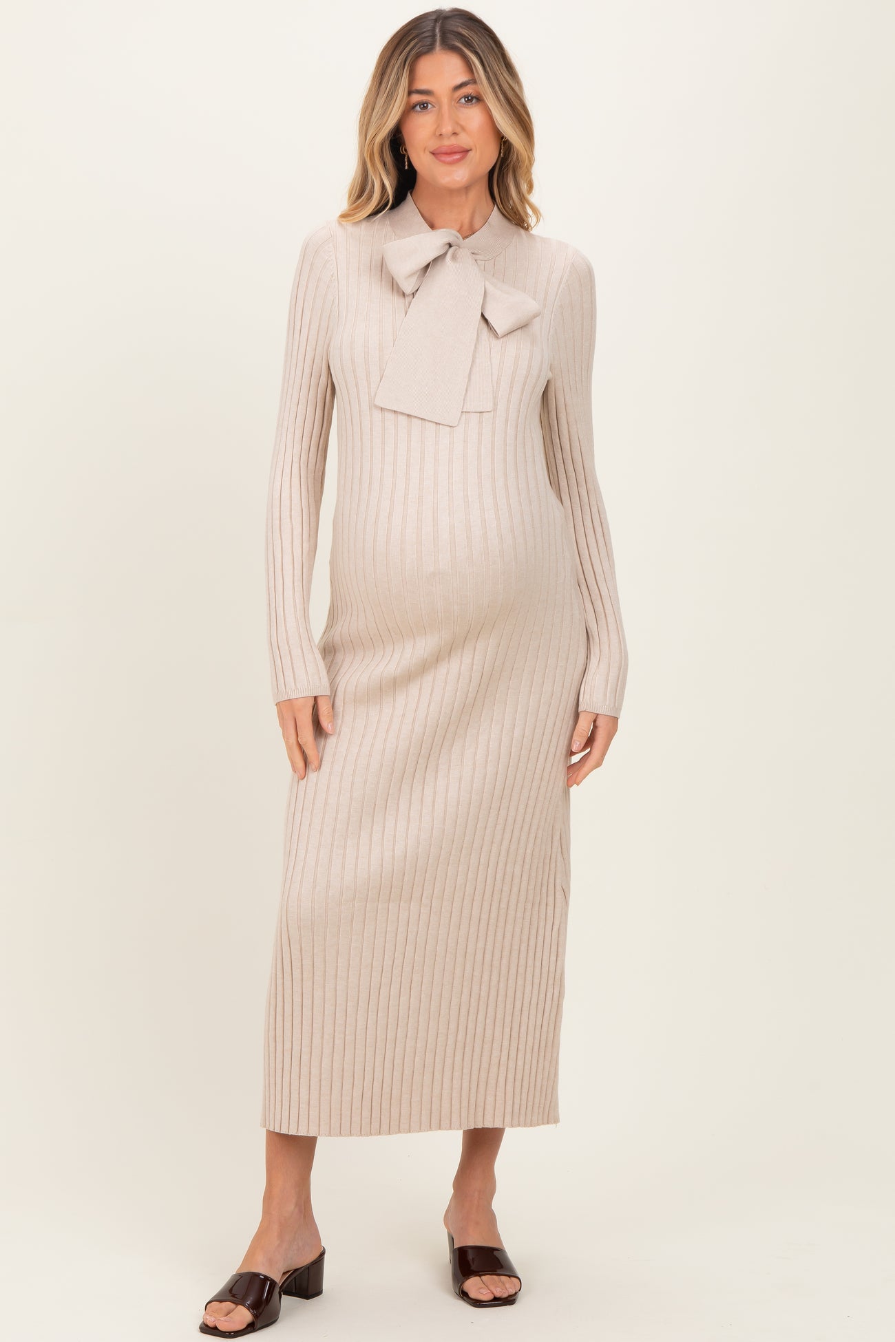woman wearing a light beige maternity sweater dress with a bow tie detail and long sleeves, showing her baby bump