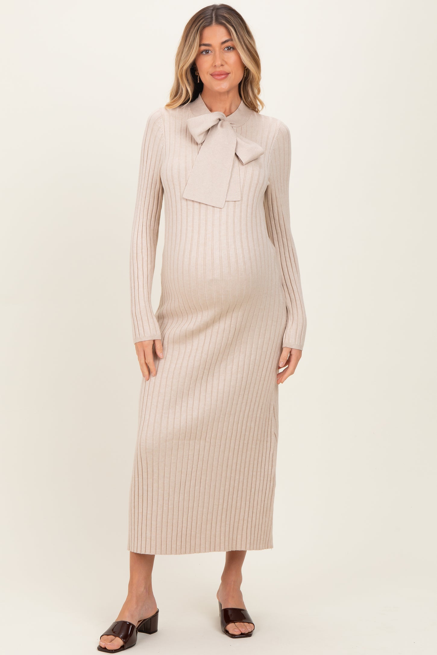 woman wearing a light beige maternity sweater dress with a bow tie detail and long sleeves, showing her baby bump