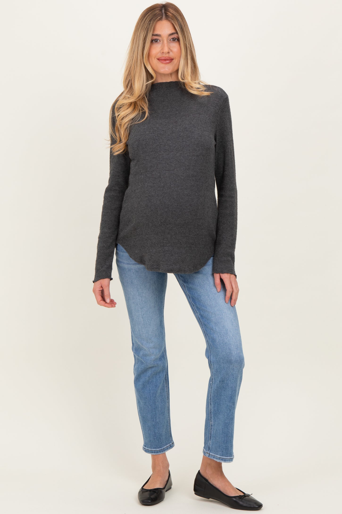 woman wearing a dark gray maternity sweater with a high neckline and blue jeans, showing a visible baby bump