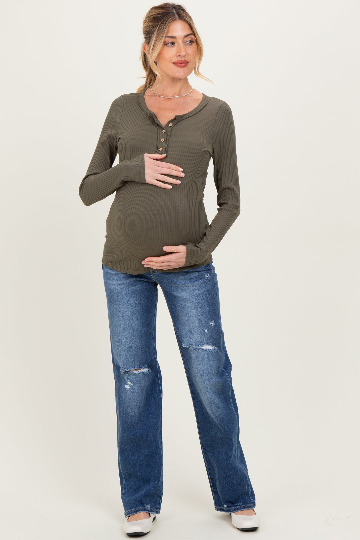 woman wearing a dark green long sleeve maternity top, holding her baby bump, paired with distressed denim jeans