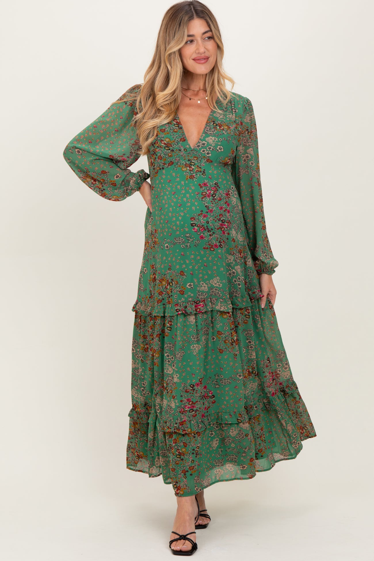 woman wearing a green floral maxi maternity dress with long sleeves and a v-neckline, smiling and standing gracefully
