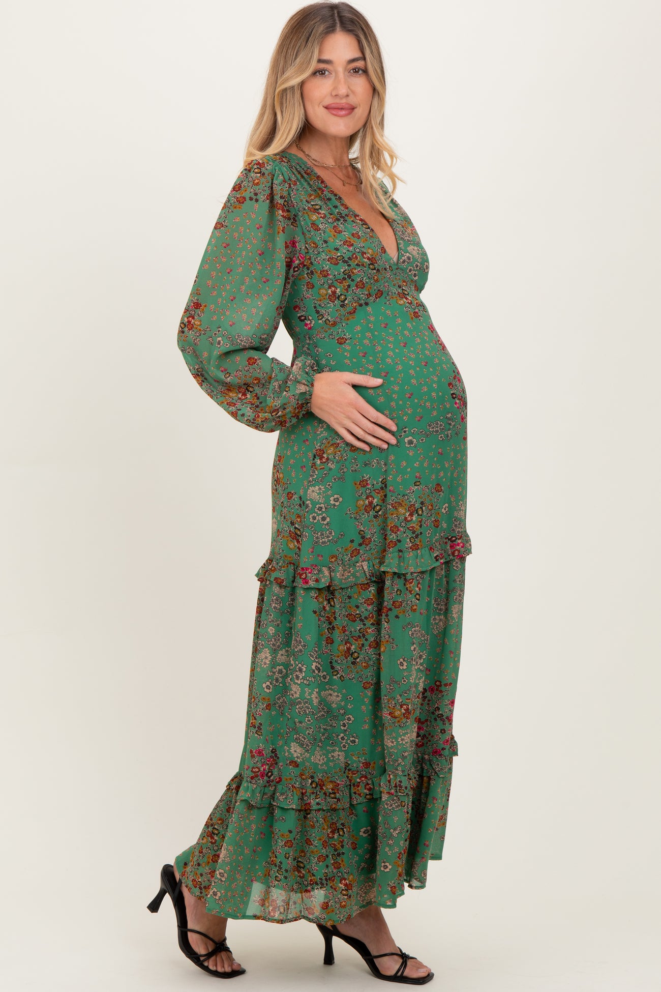 woman wearing a green floral maternity maxi dress with long sleeves and ruffles, cradling her baby bump