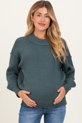 woman wearing a dark green maternity sweater, holding her baby bump with a relaxed fit and ribbed texture