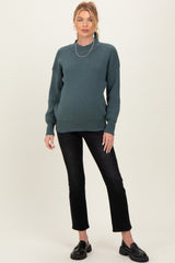 woman wearing a dark teal maternity sweater and black jeans, showing her baby bump, with a layered necklace