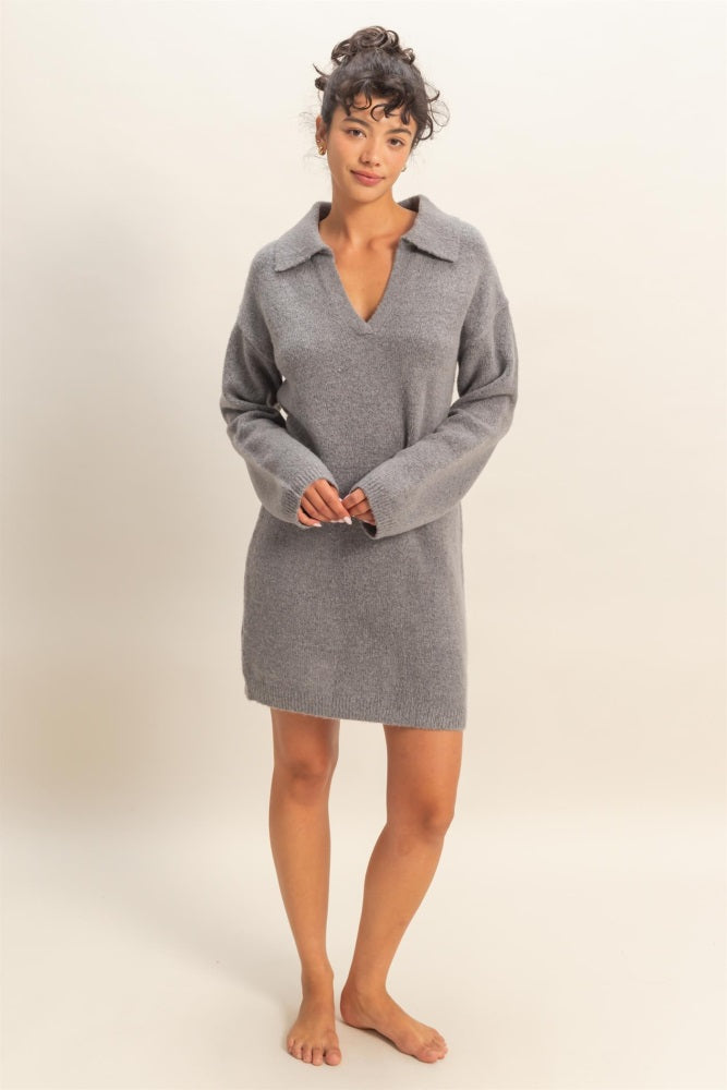 woman wearing a gray sweater dress with a v-neck and collar, standing barefoot against a neutral background
