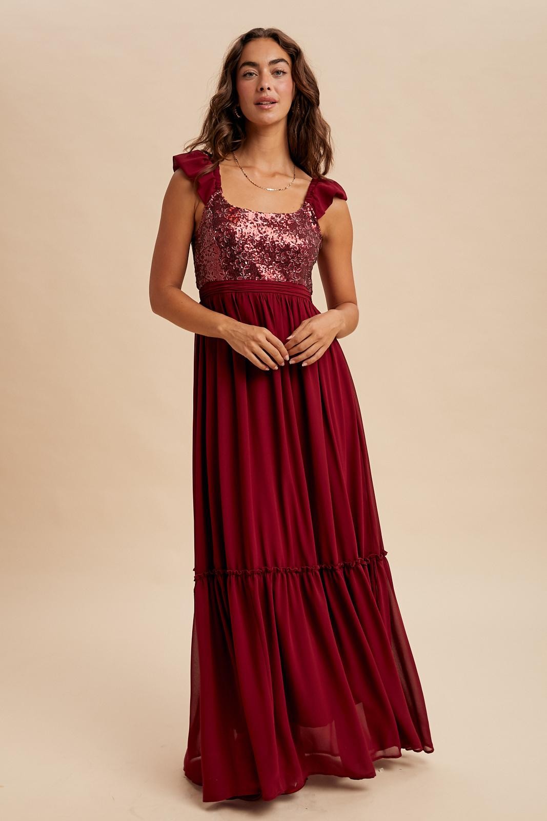 woman wearing a burgundy maxi dress with sequined bodice and ruffled sleeves, standing against a neutral background