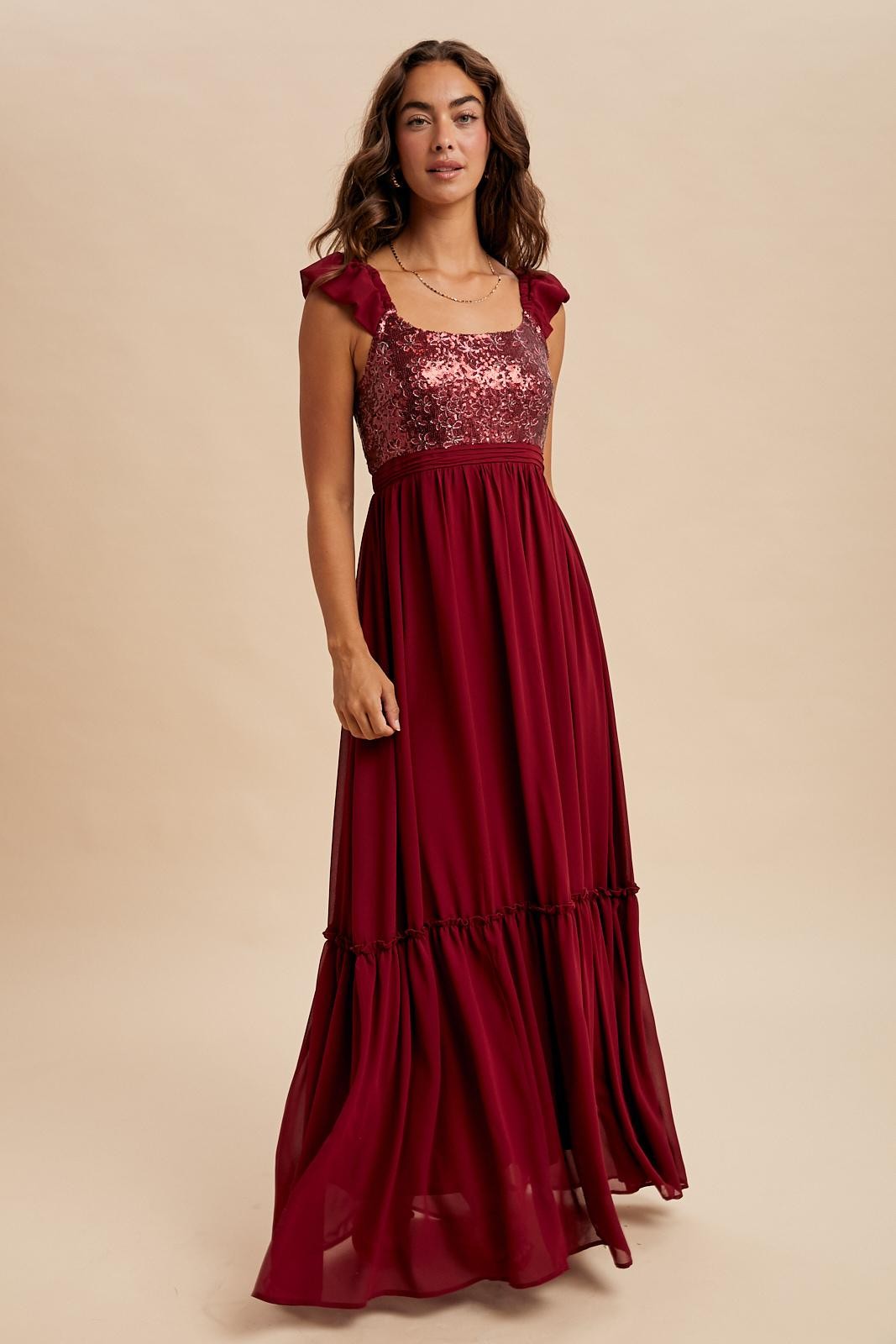 woman wearing a burgundy maxi dress with sequined bodice and ruffle straps, standing against a neutral background