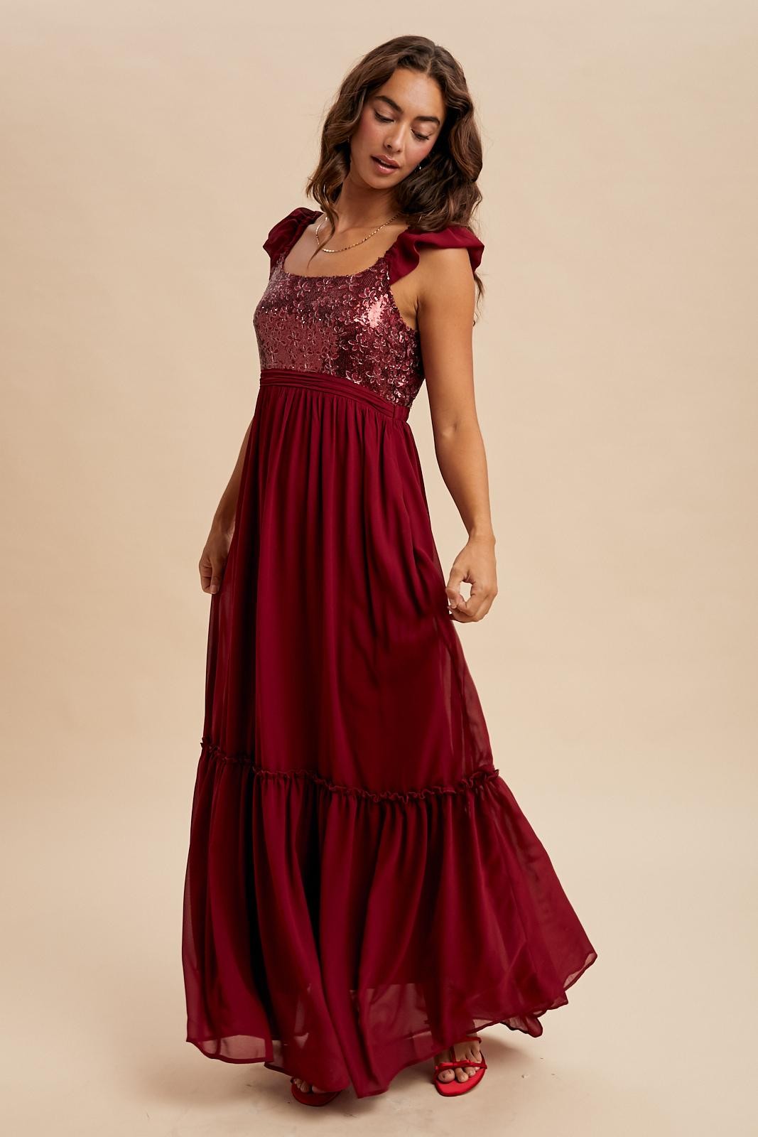 woman wearing a burgundy maxi dress with sequined bodice and ruffle sleeves, standing against a neutral background