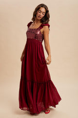 woman wearing a burgundy maxi dress with sequined bodice and ruffle sleeves, standing against a neutral background