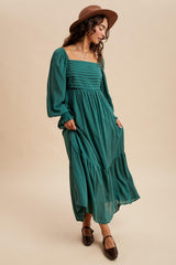 woman wearing a teal long sleeve maxi dress with gathered detailing and a flowing silhouette, accessorized with a brown hat