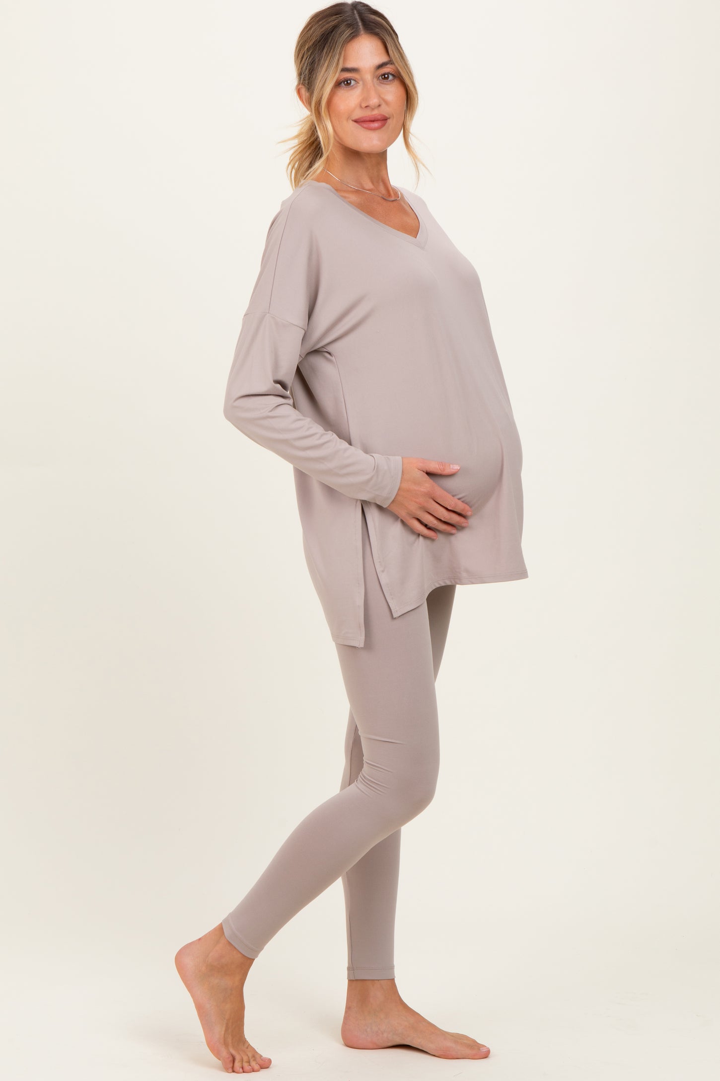 woman wearing a taupe maternity top and matching leggings, gently holding her baby bump
