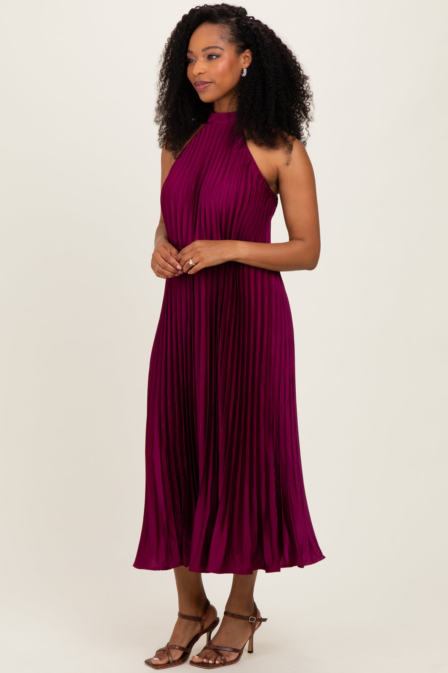 woman wearing a sleeveless pleated midi dress in deep burgundy with a halter neck and styled curls