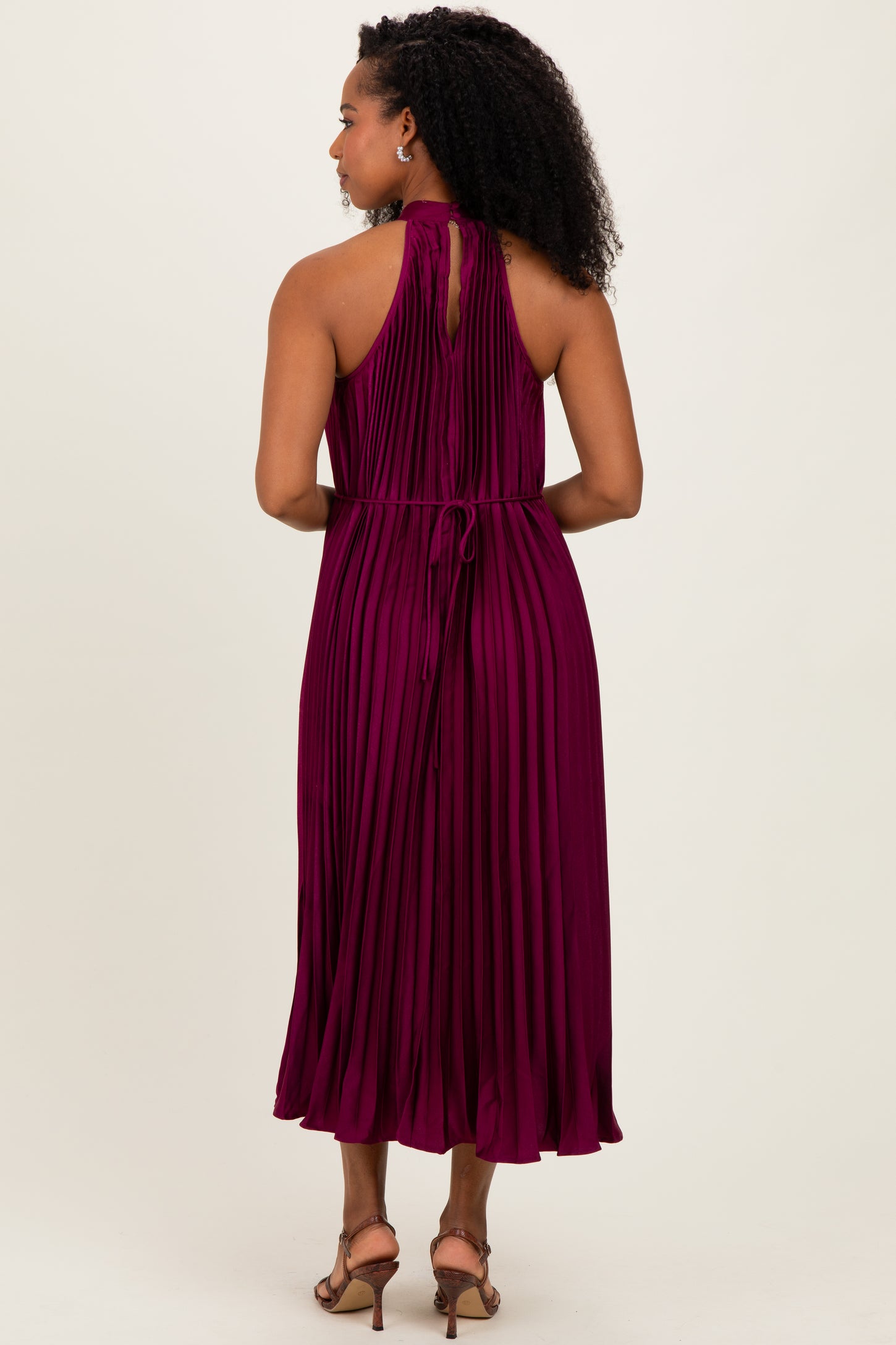 woman wearing a sleeveless pleated burgundy dress with a halter neck and back tie detail, viewed from the back