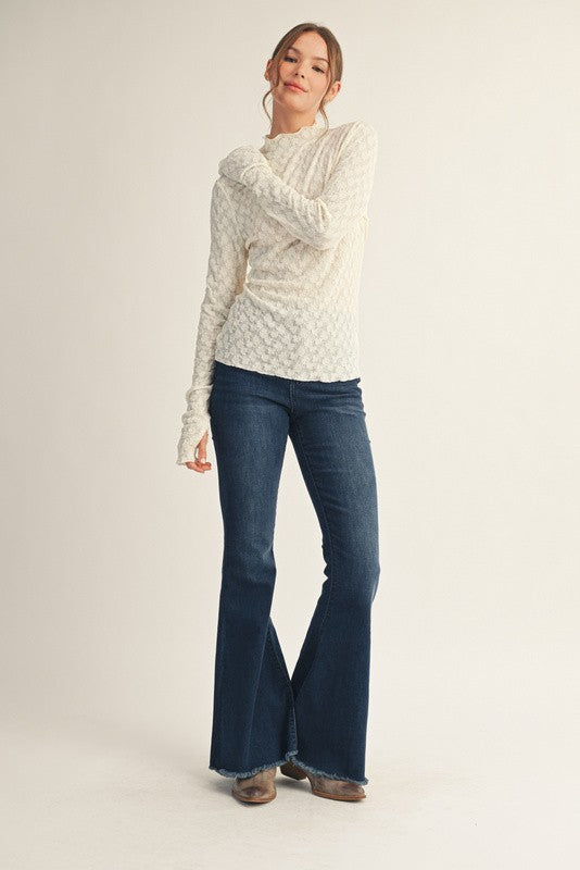 woman wearing a cream long sleeve textured sweater and dark blue flared jeans, standing with one arm crossed over her body