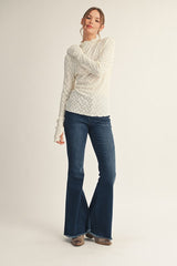 woman wearing a cream long sleeve textured sweater and dark blue flared jeans, standing with one arm crossed over her body