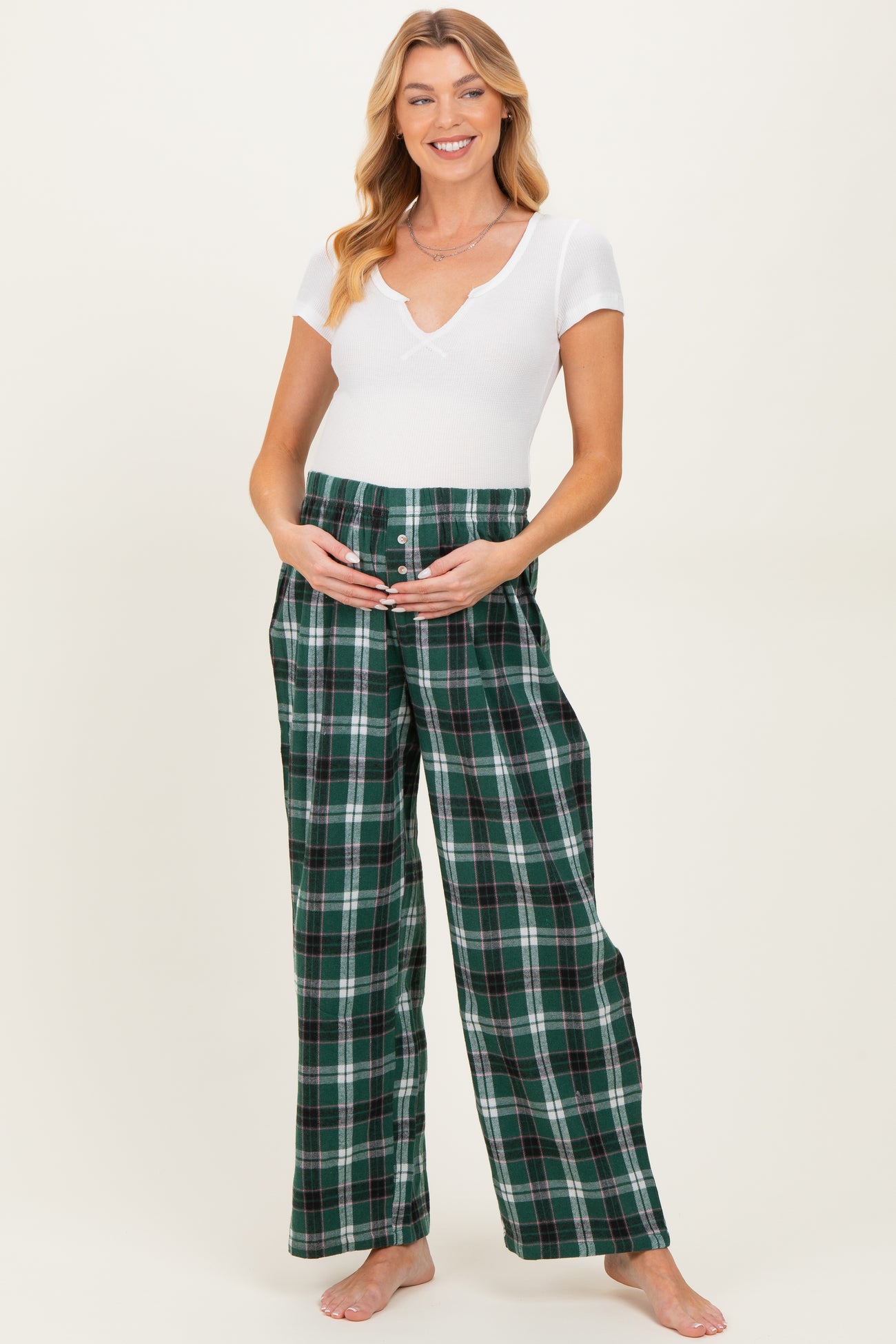 woman wearing a white short sleeve ribbed top and green plaid maternity pants, holding her baby bump