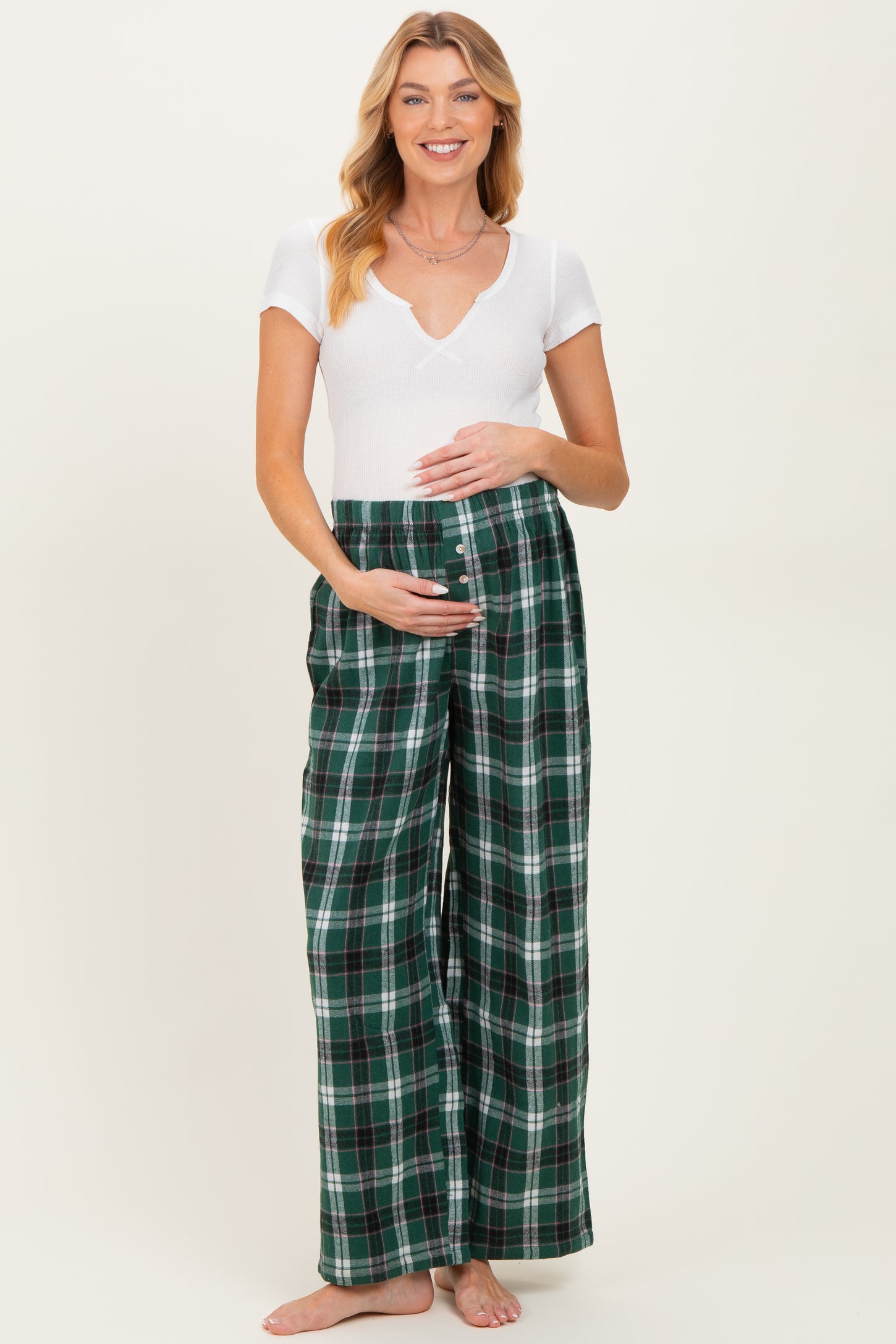 woman wearing a white short sleeve v-neck maternity top and green plaid maternity pants, holding her baby bump