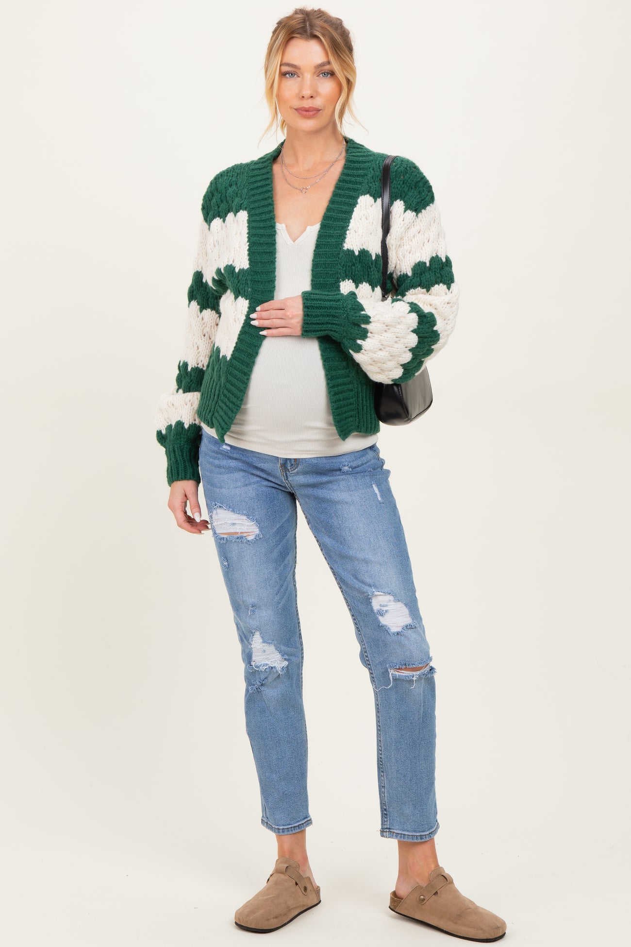 woman wearing a green and cream striped maternity cardigan and distressed jeans, gently holding her baby bump