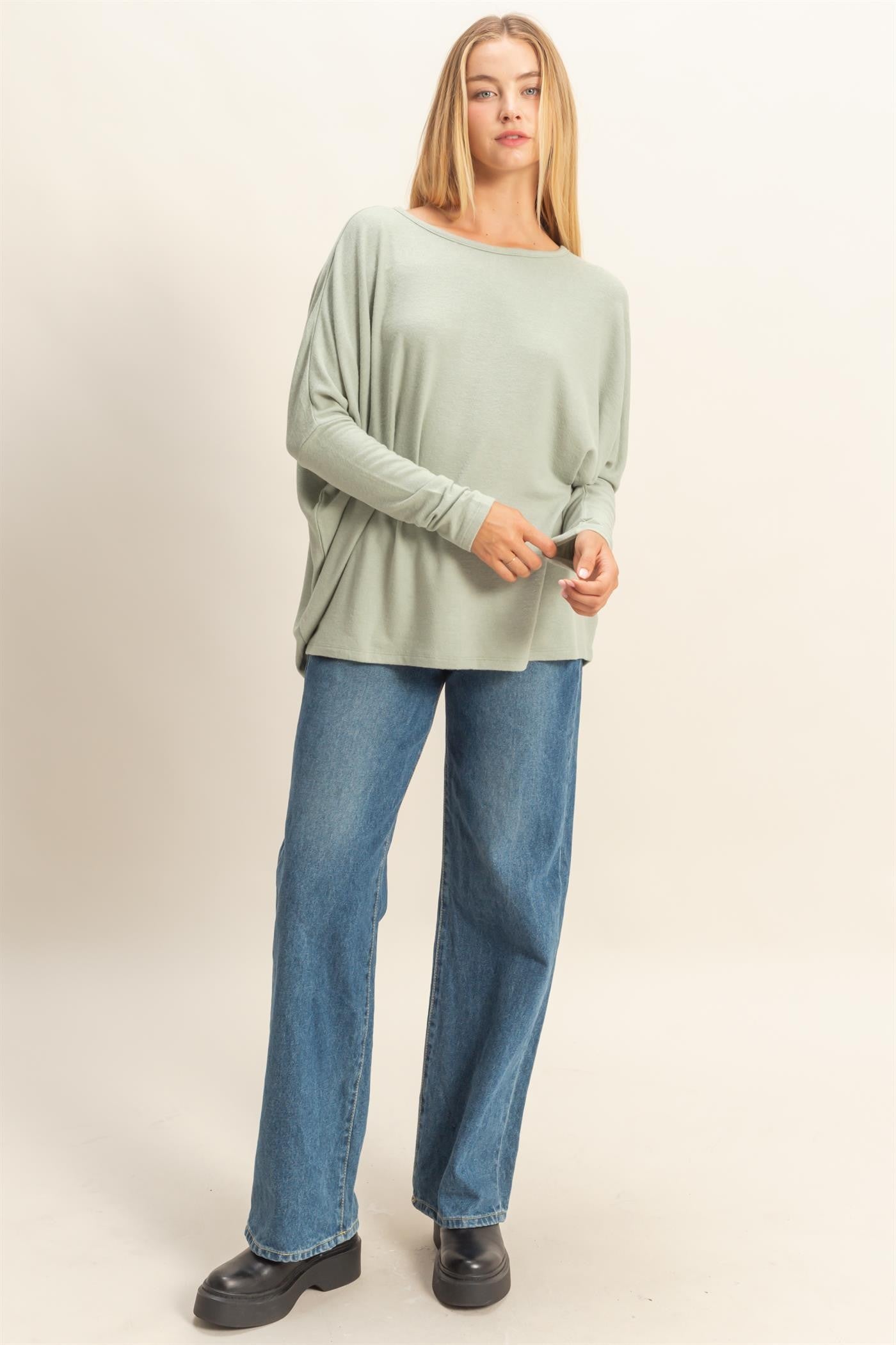 woman wearing a light green oversized long sleeve top and blue wide-leg jeans, standing with one hand on her hip