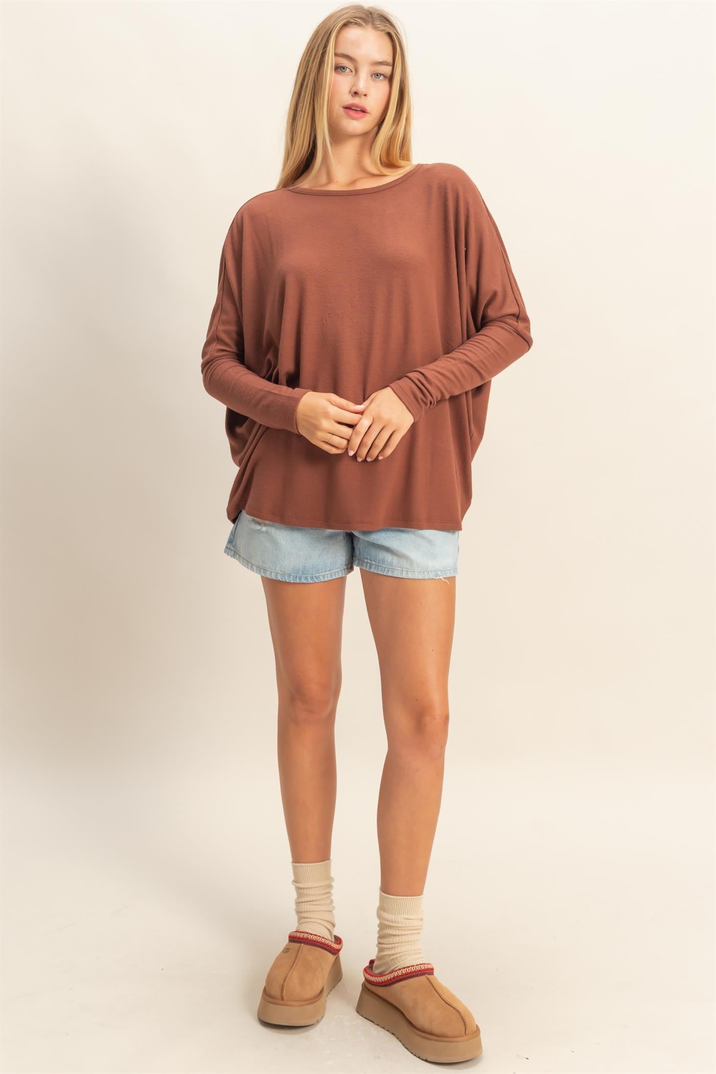 woman wearing a brown oversized long sleeve top with loose sleeves and denim shorts, standing with a relaxed pose