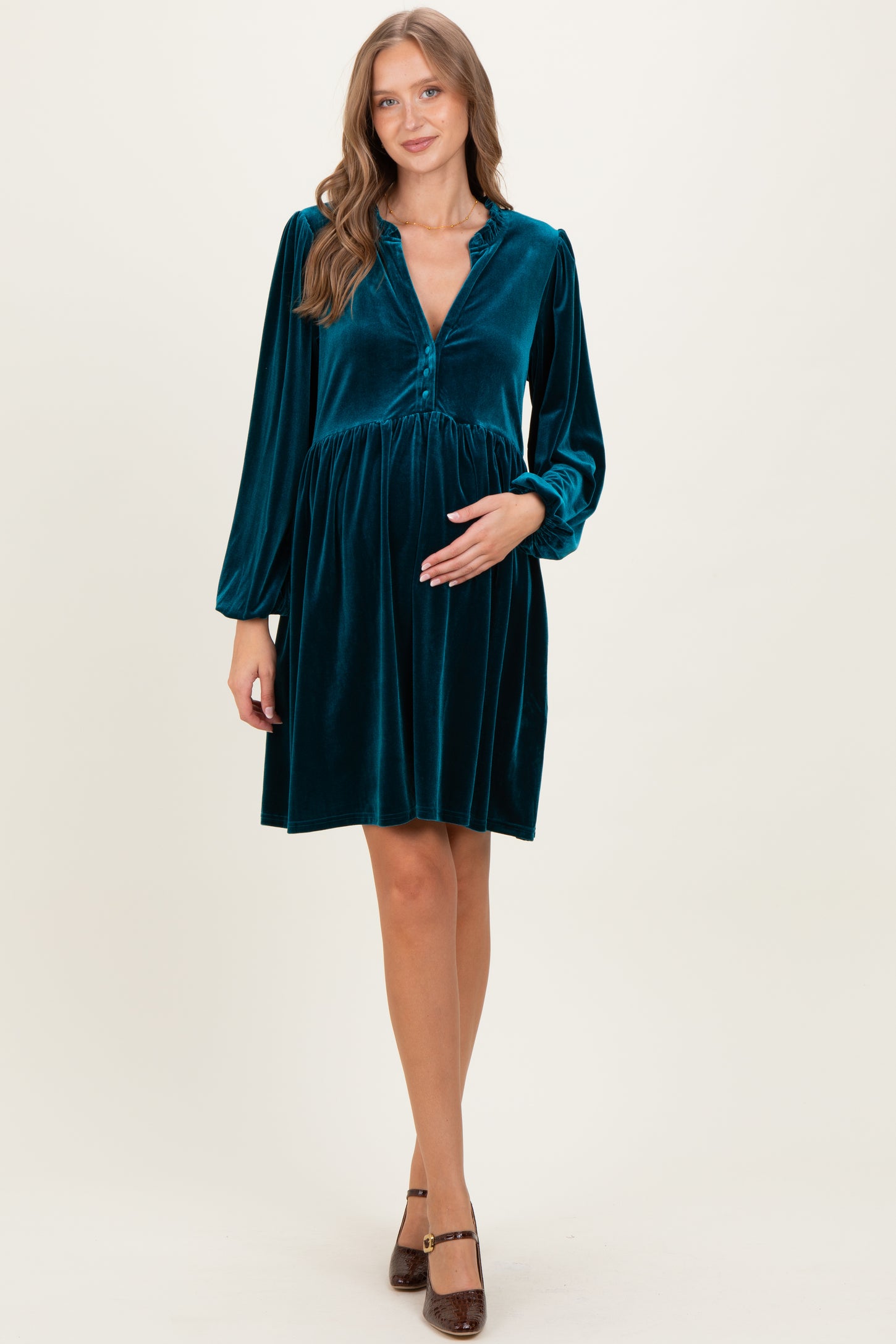 woman wearing a teal velvet maternity dress with long sleeves and a buttoned v-neck, gently cradling her baby bump