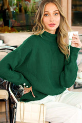 woman wearing a dark green oversized turtleneck sweater and holding a drink while sitting at a café