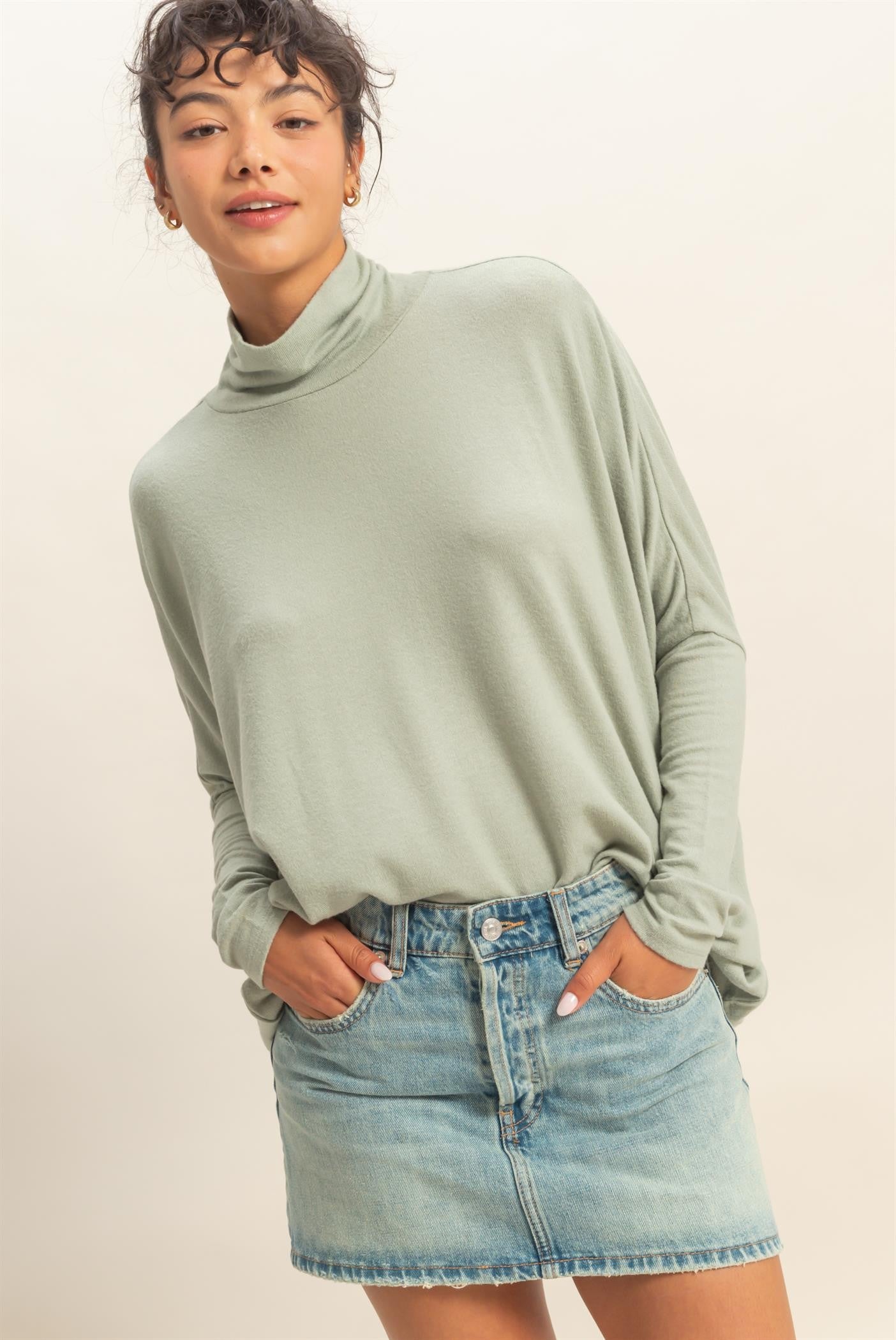 woman wearing a light green long sleeve turtleneck top and a denim mini skirt, posing with hands in pockets