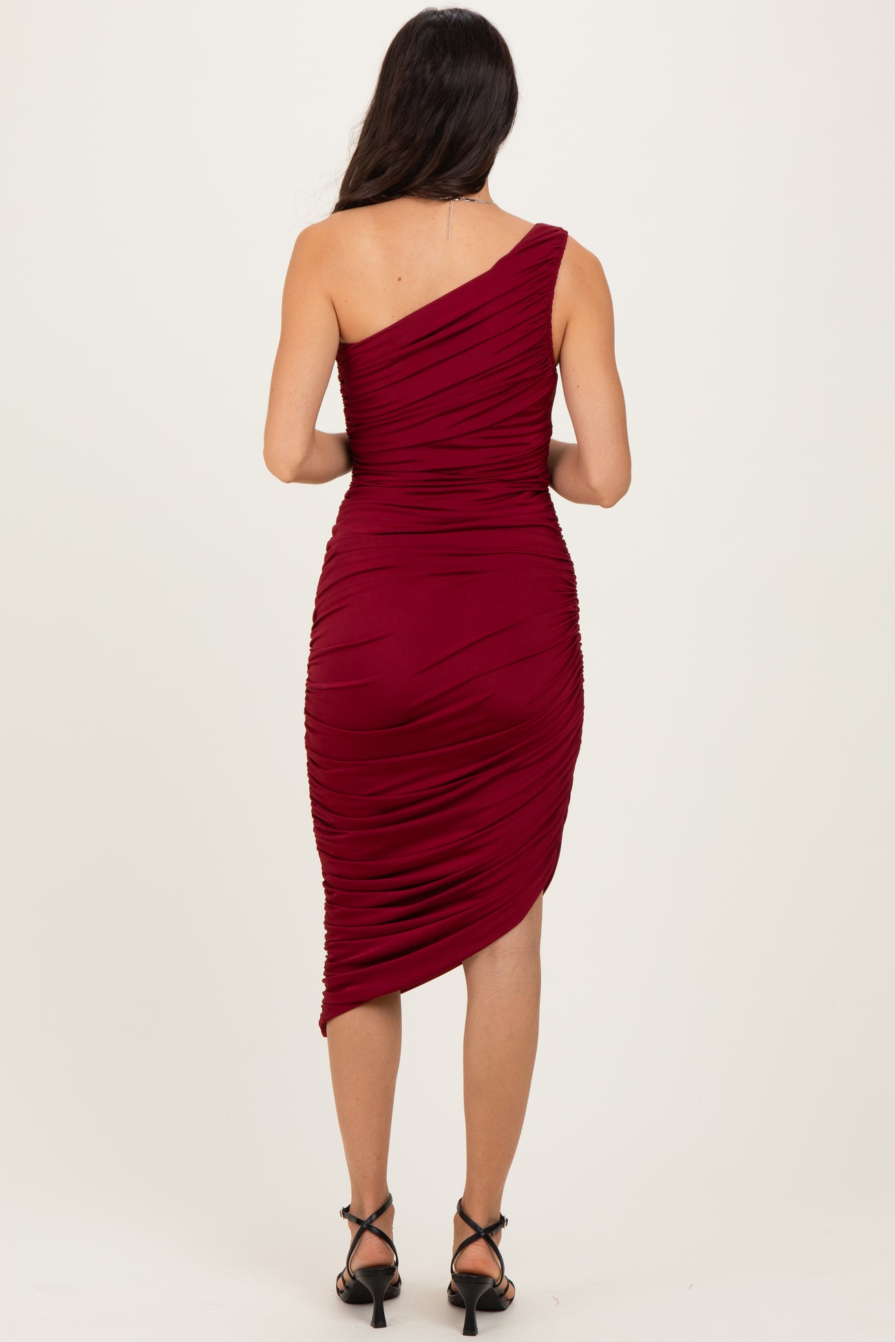 woman wearing a burgundy one-shoulder ruched midi dress, view from the back with a side slit and fitted silhouette
