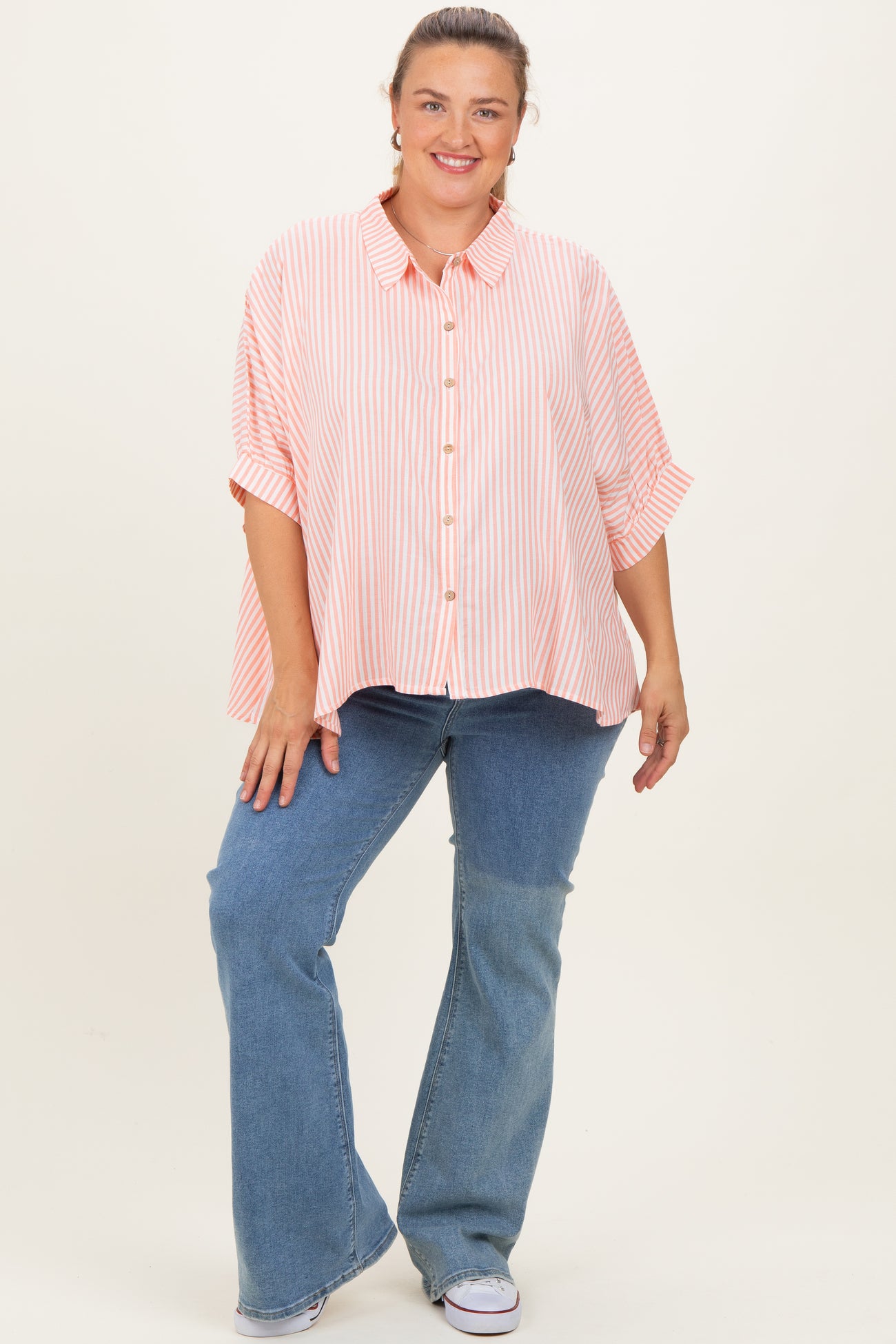 woman wearing a striped pink maternity plus shirt with buttons and rolled sleeves, paired with blue flared jeans