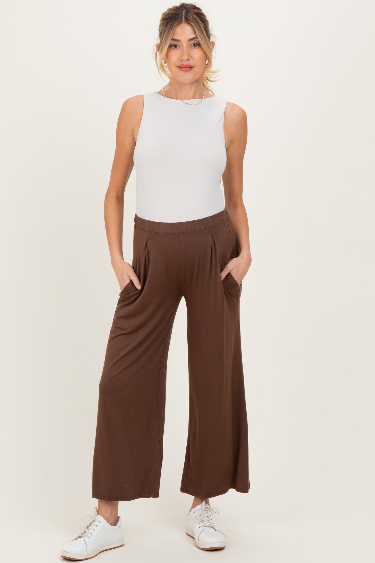 woman wearing a white sleeveless maternity top with brown wide-leg maternity pants, hands in pockets, against a neutral background