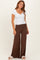 woman wearing a white ribbed short sleeve top and brown wide-leg pants, standing with one hand on her hip