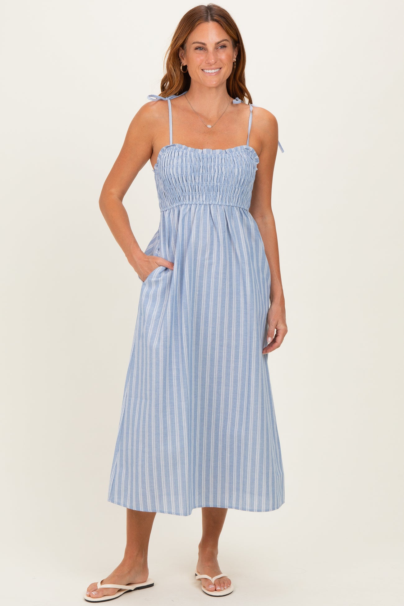 woman wearing a light blue striped maxi dress with smocked bodice and adjustable straps, posing with pockets