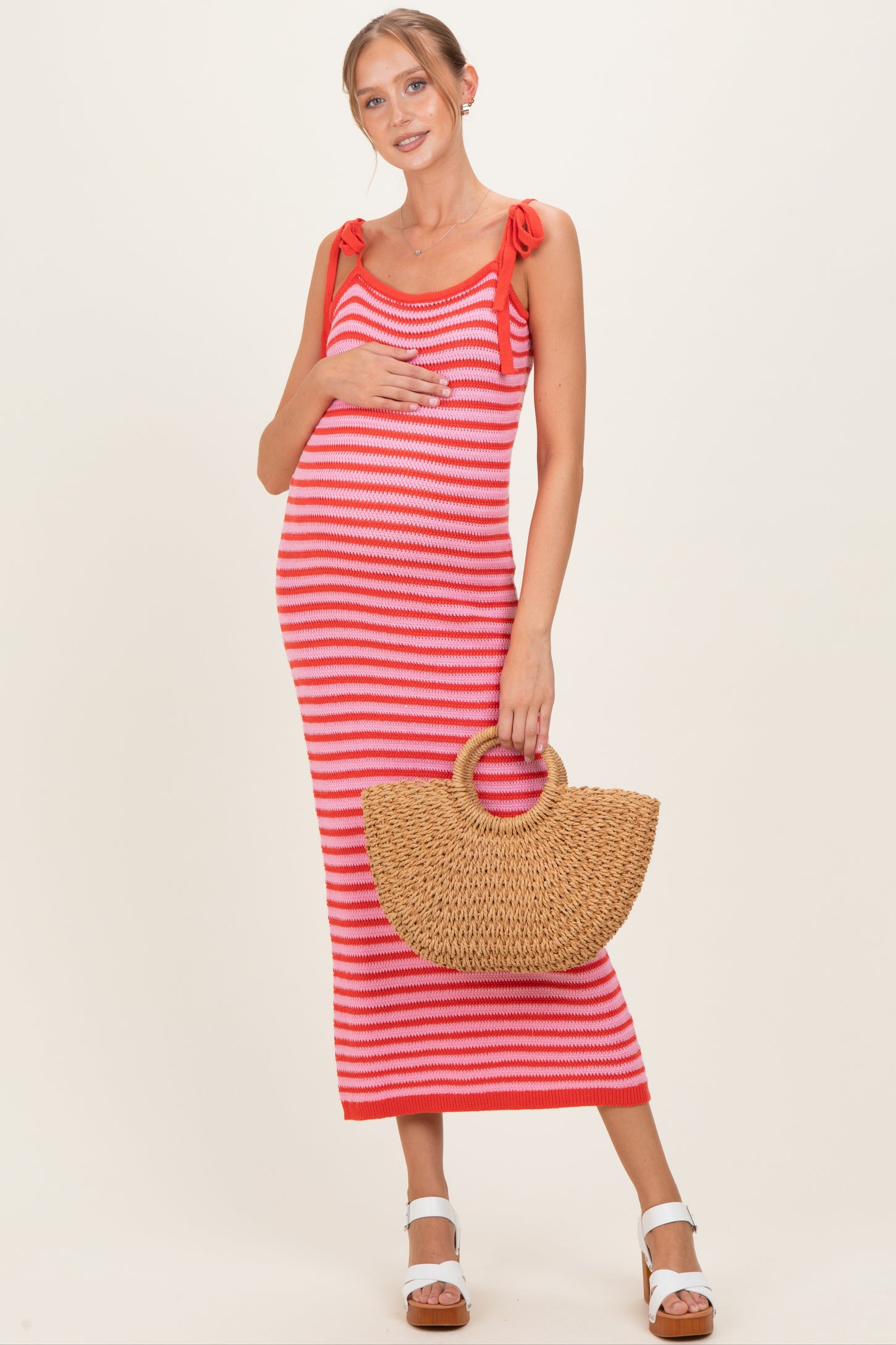 woman wearing a red and white striped maternity maxi dress with tie shoulder details and holding a straw bag, showing her baby bump