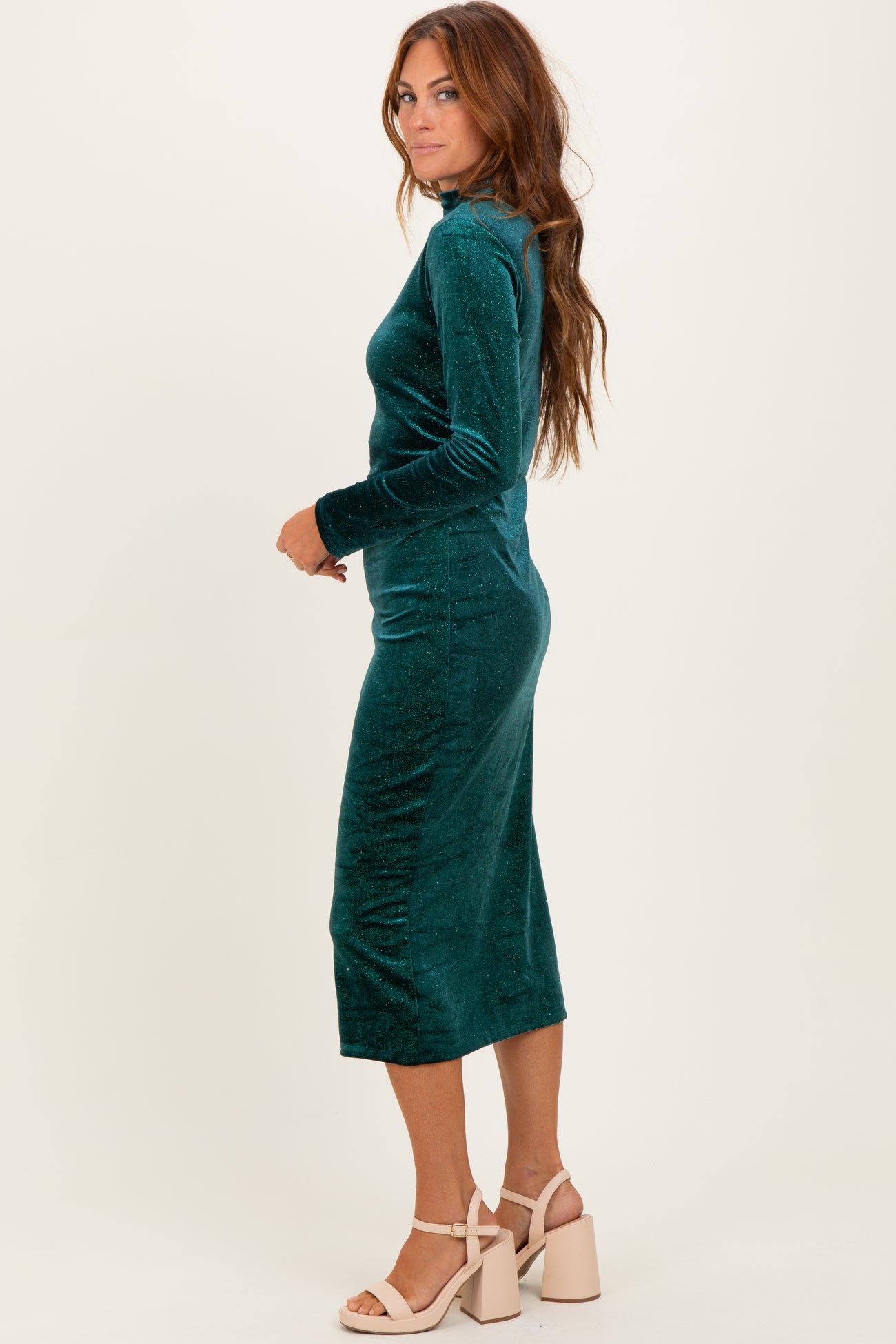woman wearing a teal velvet long sleeve midi dress with a fitted silhouette and side slit, standing sideways