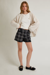 woman wearing a white fitted top, oversized beige cardigan, and black plaid shorts with frayed hem