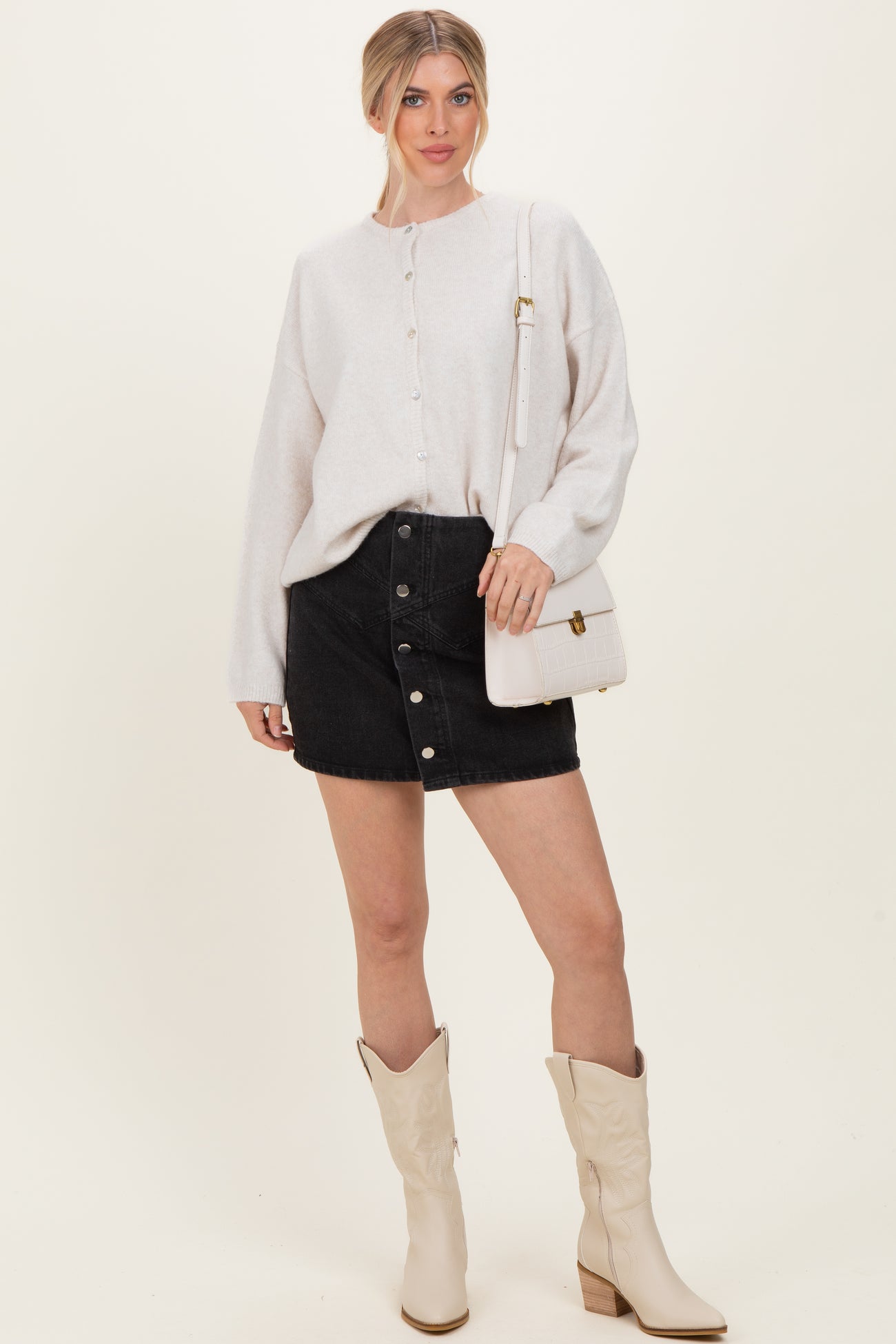 woman wearing a light gray button-up sweater, black denim button-front skirt, and cream cowboy boots, holding a white handbag