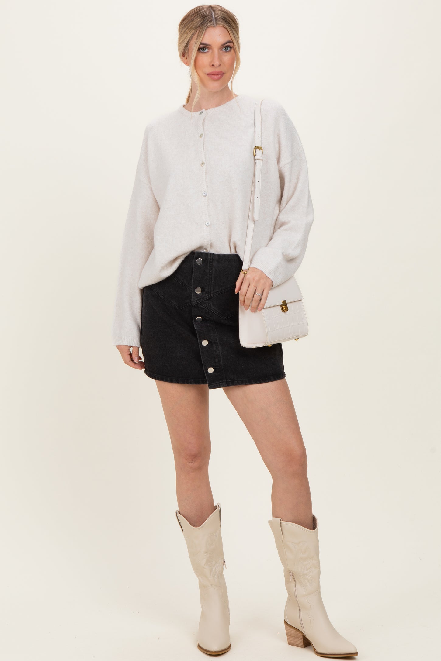 woman wearing a light gray button-up sweater, black denim button-front skirt, and cream cowboy boots, holding a white handbag