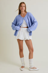 woman wearing a light purple oversized cardigan, white crop top, and white ruffled shorts, paired with white leg warmers