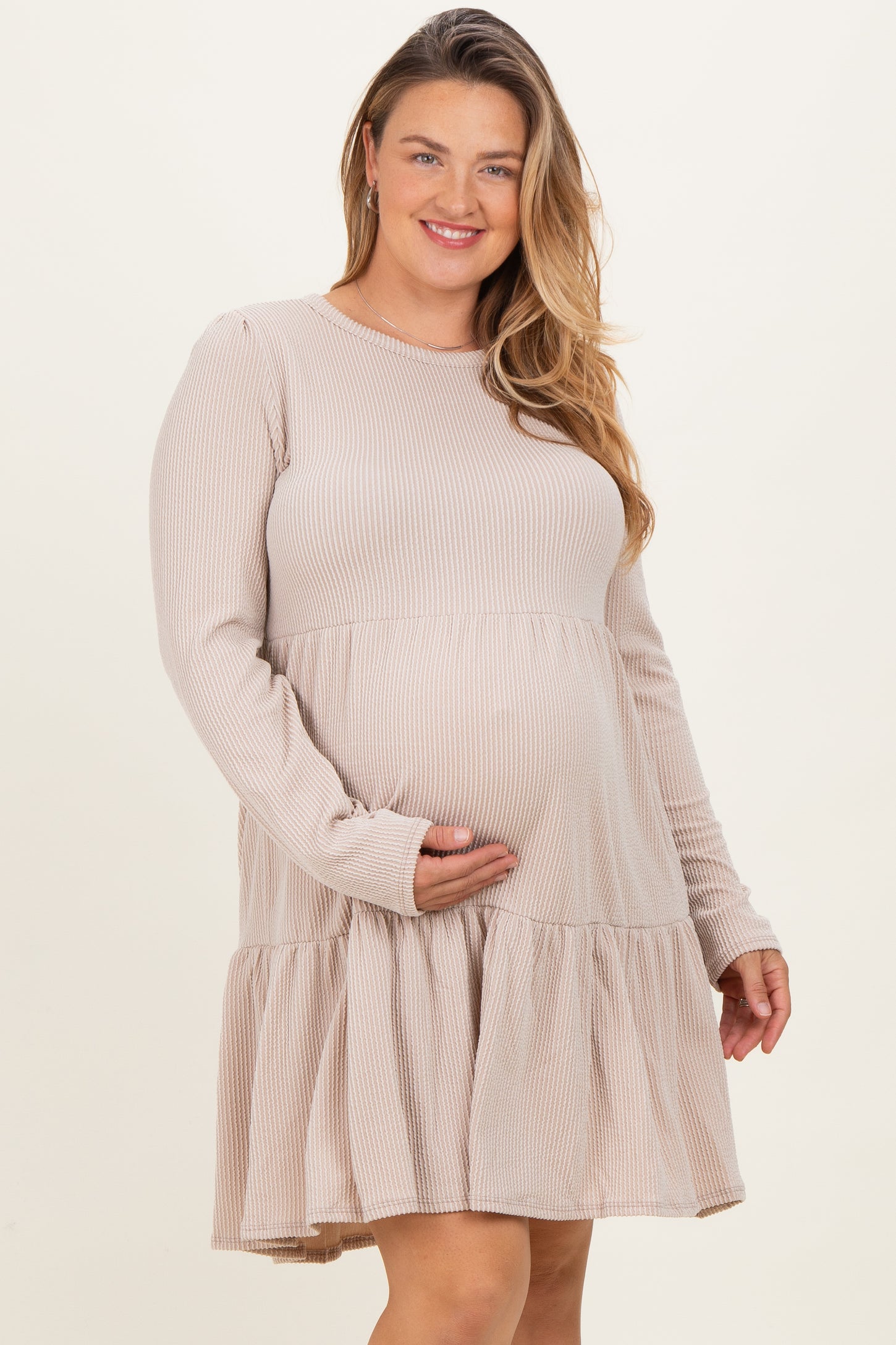 woman wearing a beige long sleeve maternity dress with a tiered skirt and holding her baby bump
