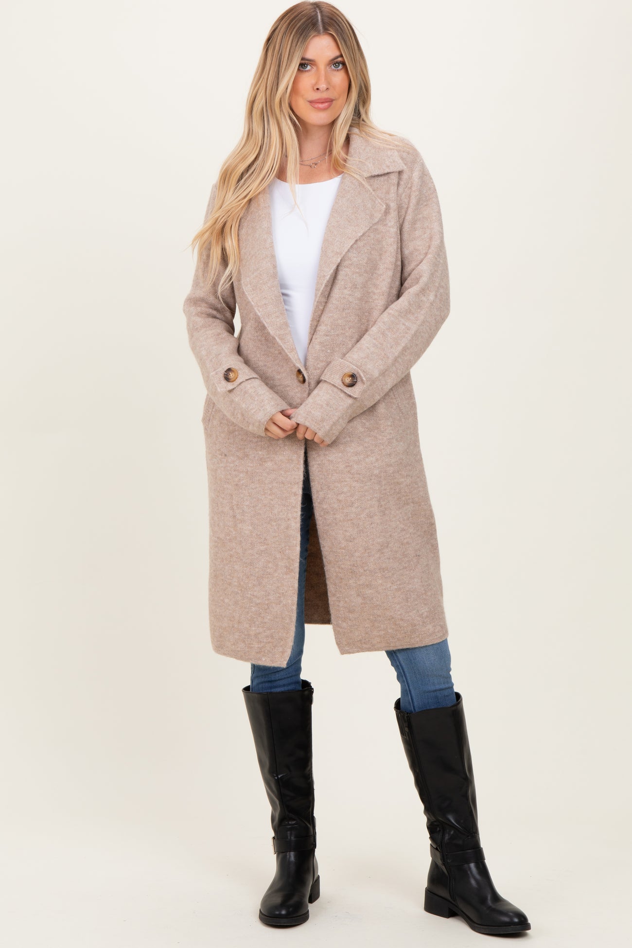 woman wearing a tan long sleeve coat with a wide collar, paired with a white top, blue jeans, and black knee-high boots