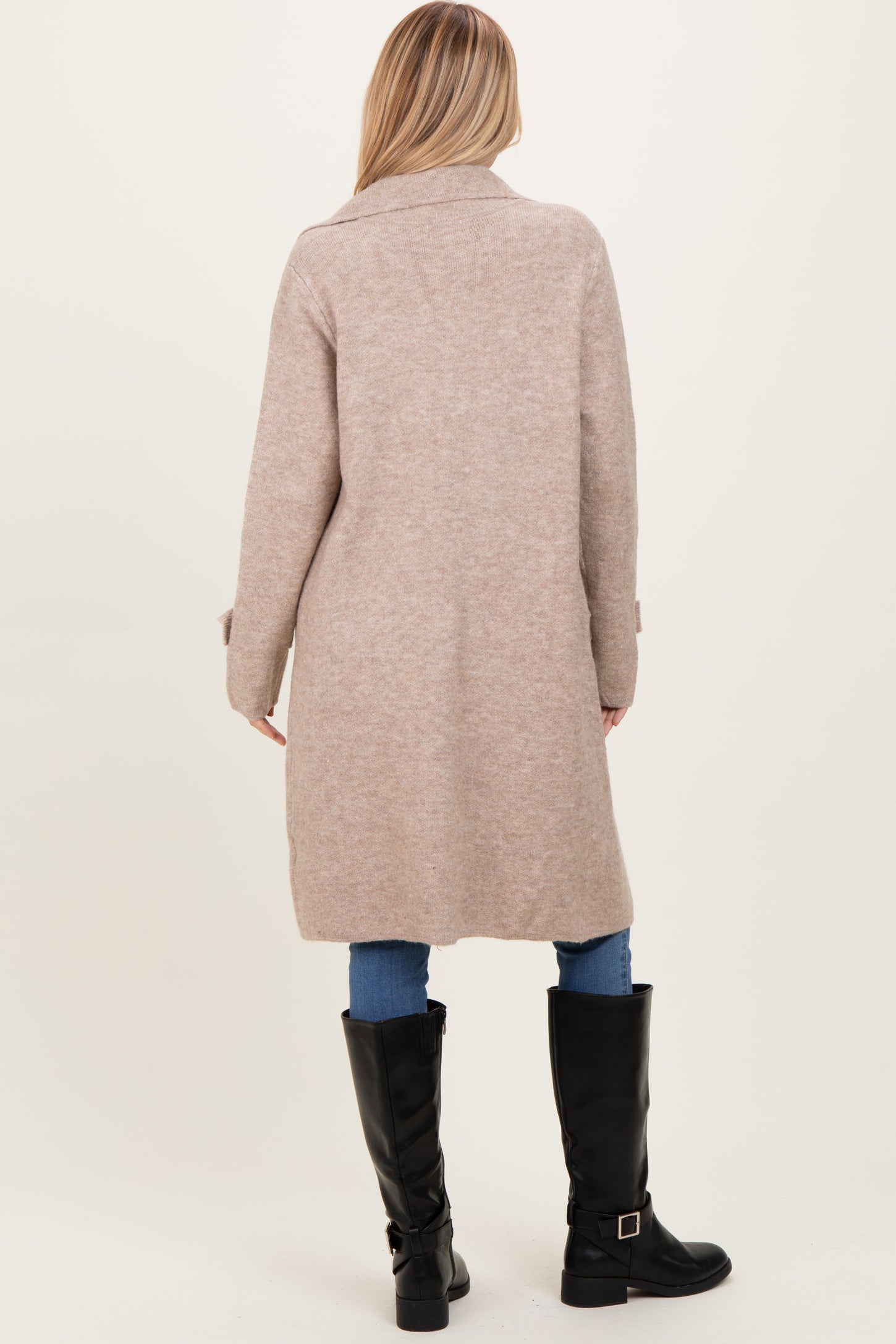woman wearing a long beige cardigan, styled with blue jeans and black knee-high boots, shown from the back