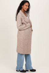 woman wearing a beige long maternity coat, holding her baby bump, paired with light blue wide-leg jeans