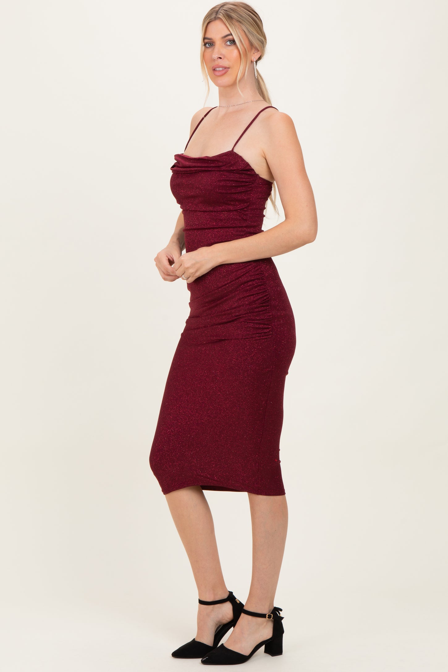 woman wearing a burgundy sparkly midi dress with ruching and thin straps, styled with black ankle-strap heels