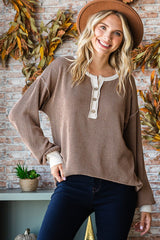 woman wearing a brown henley sweater with decorative buttons and cream detailing, smiling with a hat in a cozy setting