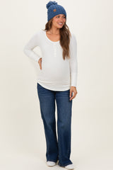 woman wearing a white maternity long sleeve henley top, flared denim jeans, and a blue beanie with a baby bump visible