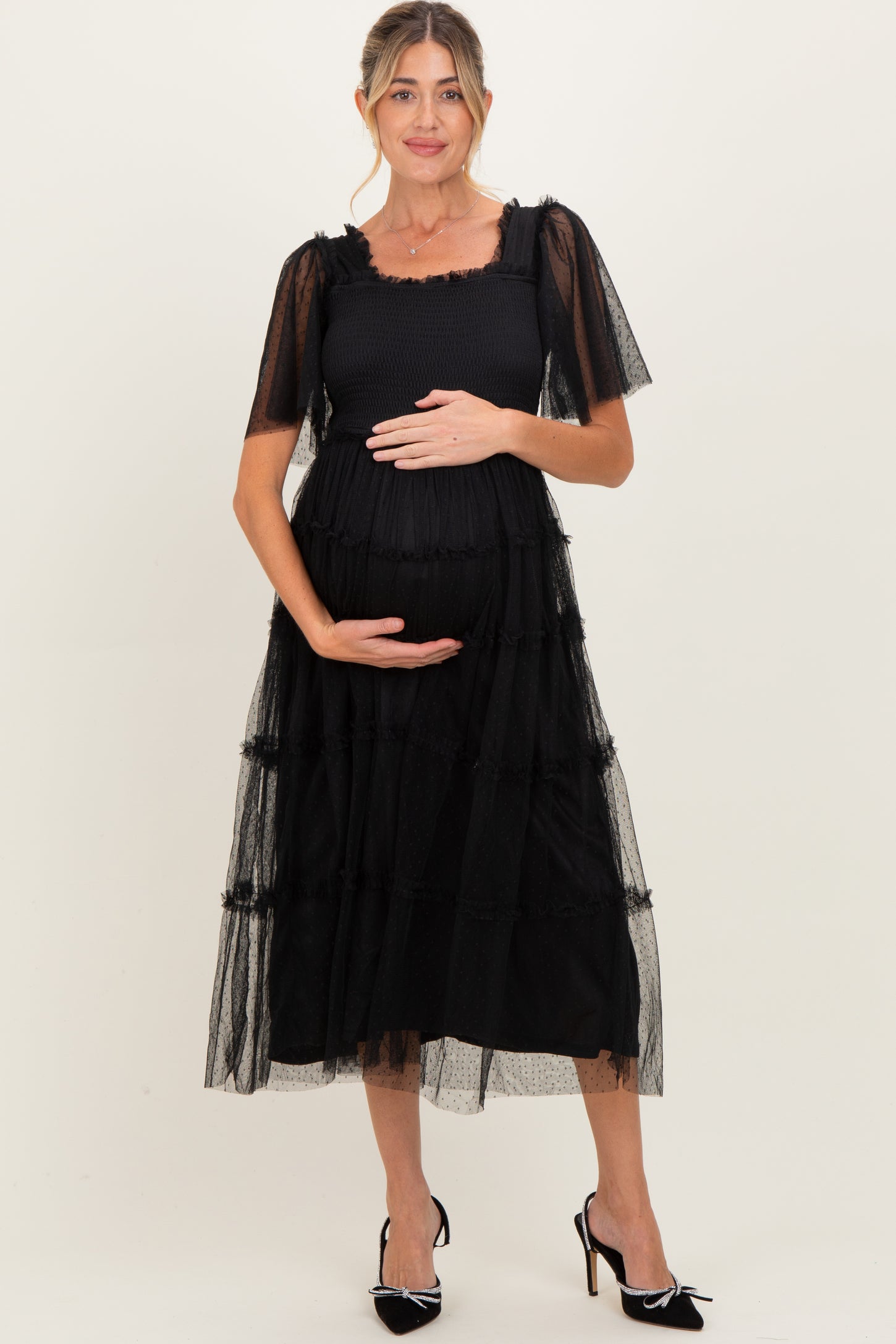 woman wearing a black maternity dress with sheer sleeves and tiered design, holding her baby bump