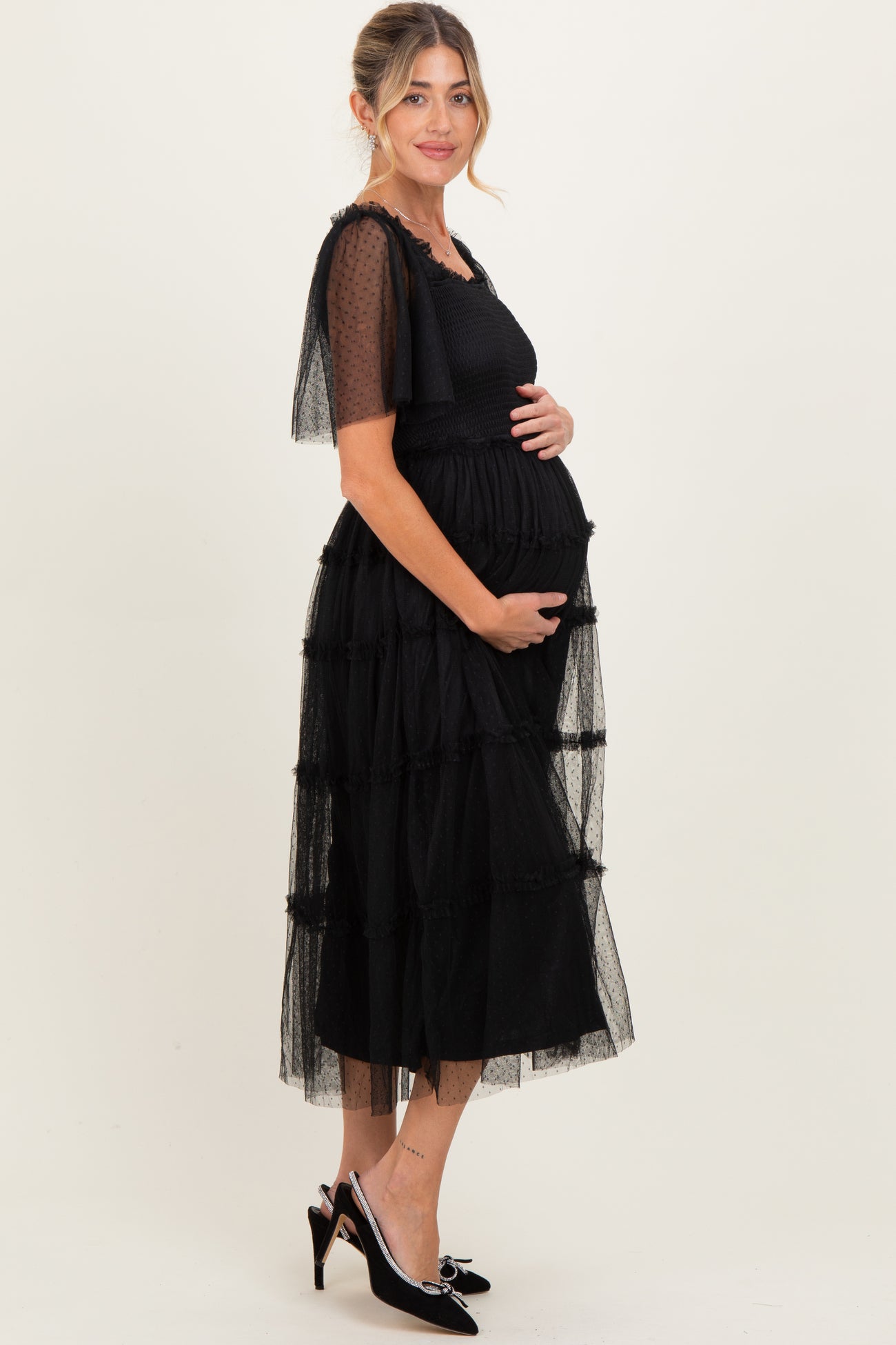 woman wearing a black tiered maternity dress with sheer puff sleeves and holding her baby bump