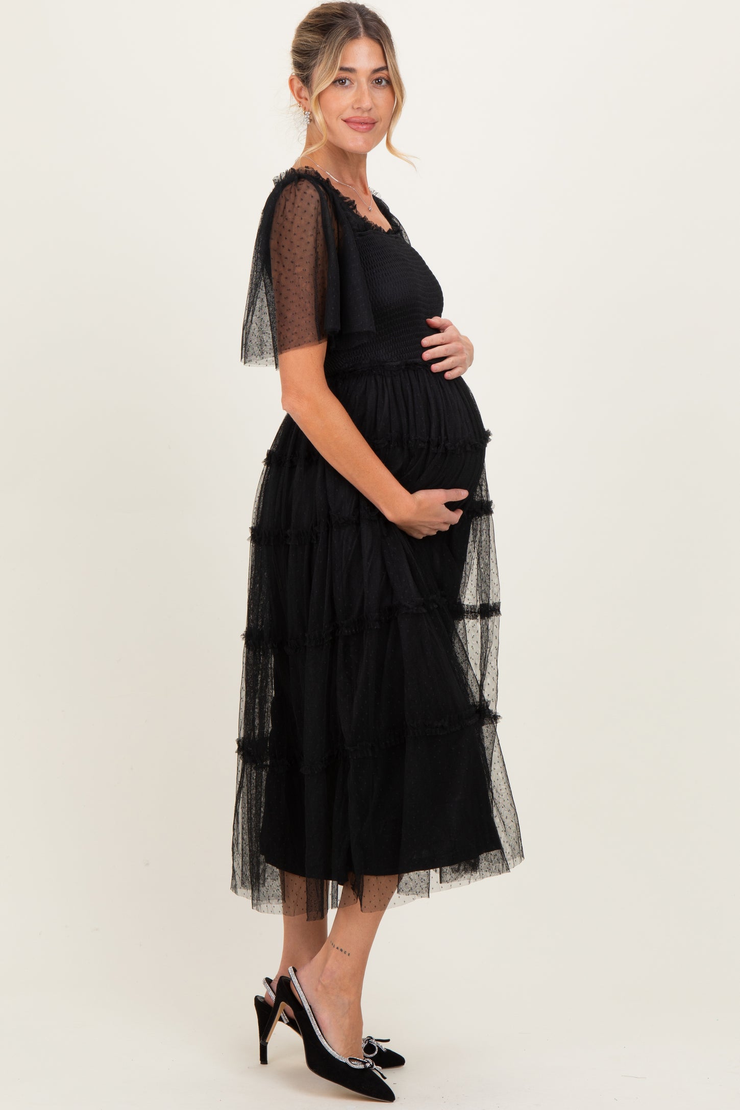 woman wearing a black tiered maternity dress with sheer puff sleeves and holding her baby bump