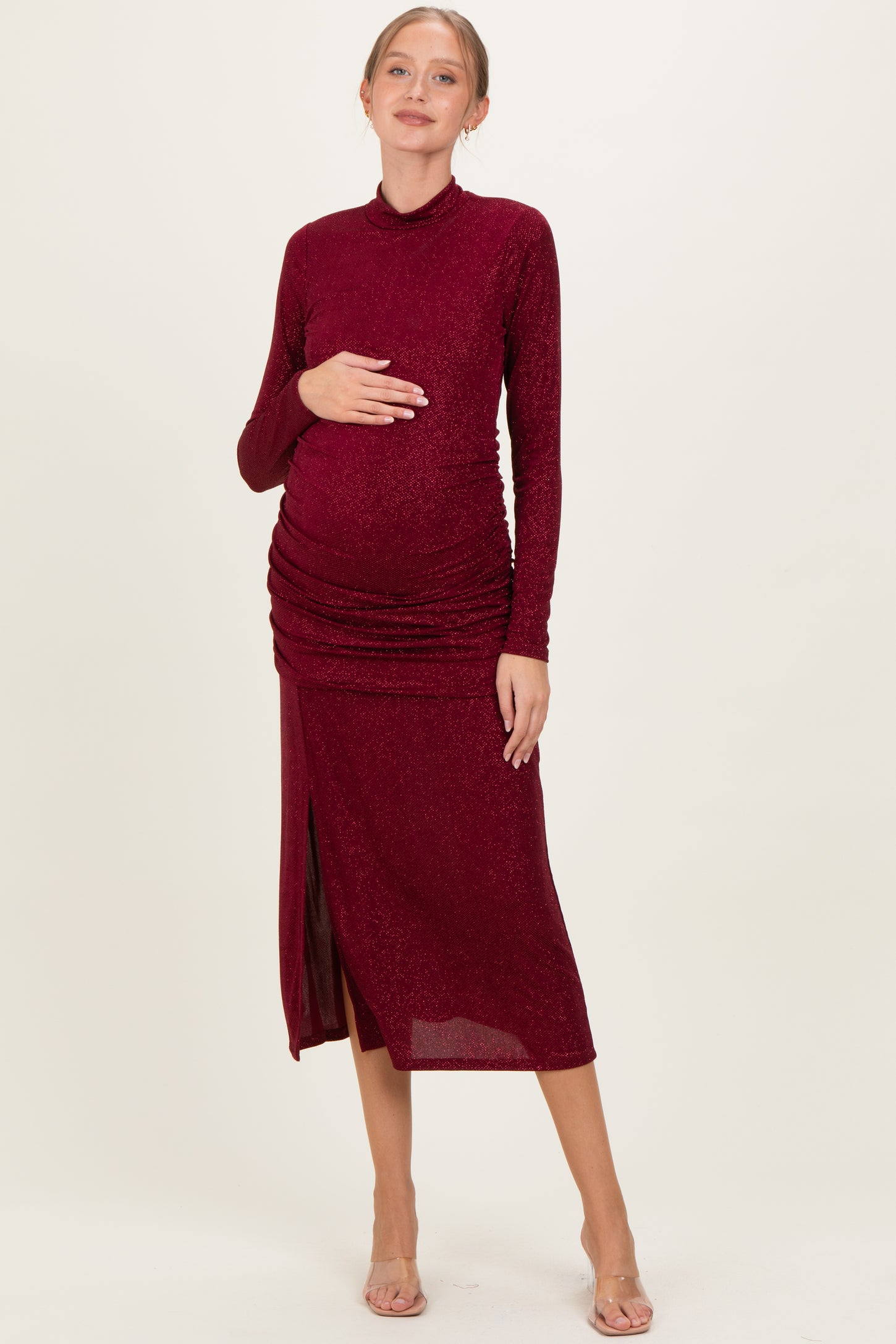 woman wearing a burgundy long sleeve maternity dress with ruched detailing and a side slit, showcasing her baby bump